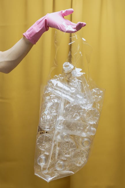 A person wearing pink rubber gloves is holding a large, clear plastic bag filled with empty transparent plastic bottles, likely consisting of water or soft drink containers. The bag is partially crumpled and the bottles inside are of various sizes, with some caps visible and others without. The background features a plain, solid yellow curtain or wall, providing a neutral backdrop that highlights the focus on the plastic waste. The scene suggests effective plastic waste collection or recycling activities, which can be associated with independent rubbish clearance services such as those offered by Rubbish Clearance Highbury, illustrating the process of separating and preparing recyclables for disposal or recycling, a common element within the context of rubbish removal and waste management tasks.