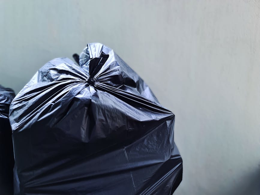 Close-up image of multiple black plastic rubbish bags filled with waste, tied securely at the top, and stacked against a plain light-colored wall. The bags are located outdoors on a paved surface, possibly a driveway or garden area, with some slightly leaning or resting on each other. The texture of the bags appears glossy and crinkled, indicating they are made of standard bin liner material. There are no visible labels or markings on the bags, which could contain household or garden waste awaiting collection. The background is minimal, with the bags occupying most of the foreground, suggesting an informal disposal or off-site rubbish collection scenario. The natural lighting highlights the reflective surface of the plastic, emphasizing the neat closure and robust packaging typical of private waste removal services like Rubbish Clearance Highbury, which provides independent rubbish clearance solutions without relying solely on local authority collections.