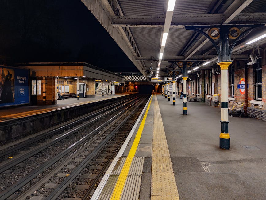 A nighttime view of a London underground train station platform, featuring multiple railway tracks running parallel along the centre. The platform is constructed from concrete with tactile yellow safety edging along the edge nearest to the tracks, designed to assist visually impaired commuters. Supporting columns with black and yellow striped bases are evenly spaced along the platform, holding up a metal canopy with an intricate Victorian-style design, painted in dark colors with yellow accents. The station's brick walls are illuminated by overhead fluorescent lighting, casting a cool, even glow across the platform and surrounding structures. On the far side, there is a small waiting area with a bench and station signage, including a roundel sign bearing the 'London Underground' logo. In the background, the tracks extend into a dark tunnel, while a single individual waiting or passing through the station is visible, sitting on a bench near the far platform edge. The scene captures a quiet period, with no trains present at the moment, emphasizing the station's spacious, well-maintained environment which is typical of UK commuter rail stations offering independent transport options for local residents.