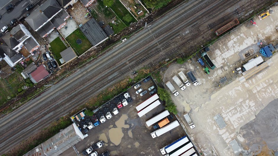 An aerial view showing a commercial and residential area intersected by railway tracks running diagonally across the image from top left to bottom right. To the top left, there are numerous small houses with pitched roofs, some with gardens and driveways, and a variety of items such as vehicles and outdoor furniture visible. The railway tracks, with visible metal rails and wooden sleepers, separate the residential area from a large paved lot at the bottom right. This lot contains several white vans, trucks, and trailers parked in an orderly manner, along with some smaller vehicles. Puddles of water are scattered across the muddy and gravelly surface of the lot, indicating recent rain. Near the bottom right corner, construction materials and equipment, including blue plastic containers, pallets, and landscaping elements, are positioned on a concrete surface. The environment appears to be a mix of urban infrastructure and utility storage, with some greenery and scattered debris, suggesting active use for waste removal or logistics services carried out by Rubbish Clearance Highbury in this setting.