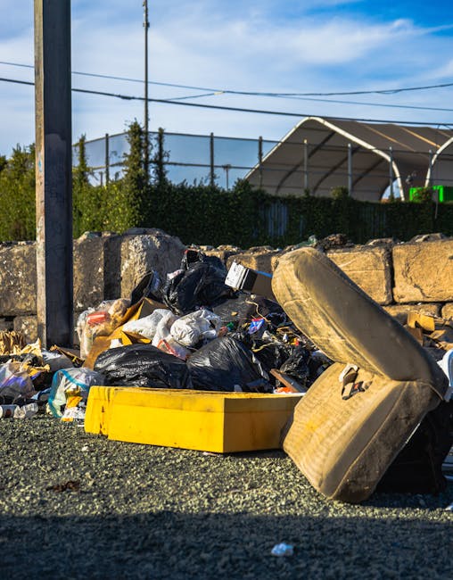 A collection of discarded rubbish piled on a gravel surface outdoors, including black plastic garbage bags, a yellow plastic container, and what appears to be an old, worn tyre or cushion leaning against the debris. Behind the pile, there is a low stone retaining wall made of irregularly shaped rocks, with some greenery such as small bushes or trees visible beyond it. To the left, part of a metal pole and electrical wires are seen, suggesting the scene is along a roadside or public space. In the background, a large, curved canopy structure with a metallic frame and semi-transparent covering is partially visible, set against a partly cloudy sky with blue tones. The overall scene indicates a case of unmanaged waste, which could be associated with alternative or private rubbish collection services like those provided by Rubbish Clearance Highbury, especially in contexts of difficult access or non-standard disposal areas.
