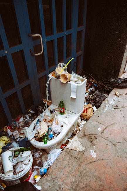A large pile of black rubbish bags, some partially torn or bulging, positioned in front of a red metal door set into a beige, textured external wall. The bags are filled with assorted waste, with some open packaging and loose debris visible beneath. To the left of the pile, there is a discarded piece of cardboard and a wooden pallet, indicating informal waste collection or clearance from a flat or alleyway. The red door features graffiti, a white sign stating 'NO MONEY' and 'Keep clear' in blue, along with another smaller faded sign, all affixed directly onto the door. The surrounding environment suggests an urban setting, possibly a back alley or service area, where private rubbish disposal might occur outside regular municipal services. The muted lighting emphasizes the utilitarian scene, aligned with the context of alternative rubbish removal and on-site clearance services offered by companies like Rubbish Clearance Highbury.