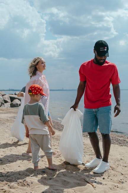 A group of three people, consisting of a man and two children, are engaged in beach clean-up activities along a sandy shoreline. The man, positioned on the right, is wearing a red T-shirt, light blue denim shorts, white sneakers, and a black cap with a red and white logo, and he is holding a large white plastic bag in his right hand, appearing to collect debris. To his left, a young boy wearing a grey T-shirt, beige pants, and a red cap with a playful pattern is observing the man, with a white plastic bag hanging from his shoulder. Behind them, a woman with curly blonde hair, dressed in a light pink jacket, stands facing the water, watching the children. In the background, the shoreline extends towards a body of water, with large rocks visible on the left and an overcast sky with scattered clouds overhead, providing natural but diffused lighting. The scene depicts an outdoor environment focused on community effort in waste collection, highlighting private or independent rubbish removal activities instead of official beach cleanup services, in line with rubbish clearance services offered by companies like Rubbish Clearance Highbury.
