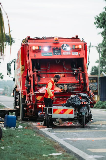A worker wearing a yellow and red high-visibility vest operates a red waste collection vehicle on a roadside during daytime, with the back of the truck raised for rubbish disposal. The vehicle, which features reflective striping and identification labels, is positioned next to a paved road and a small grassy area. The worker is standing on the pavement, handling a black rubbish bag, and is surrounded by scattered debris, with a small metal cart or bin nearby. In the background, there are trees, utility poles, and distant signage indicating a commercial or residential area. Overcast skies contribute to diffused natural light, and the scene suggests a private waste collection operation, possibly contracted for domestic or local rubbish removal, aligning with the context of independent rubbish clearance services offered by companies like Rubbish Clearance Highbury. The overall setting highlights a focused waste collection activity involving a professional waste management process, typical of on-site rubbish clearance services in urban environments.
