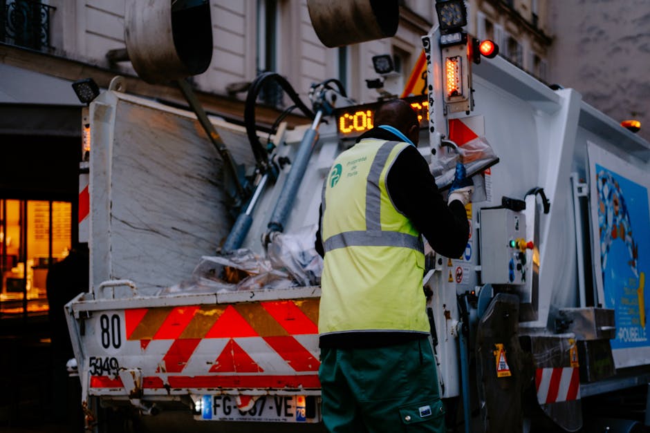 A worker wearing a high-visibility yellow vest and dark clothing is operating a waste collection vehicle, standing on the right side, with their back facing the camera. The vehicle has an open-top section filled with various types of rubbish, including paper, plastic, and other debris, accumulated near the back of the truck. The truck's side is white with red and orange reflective markings, and multiple hydraulic or mechanical components are visible, used for lifting and loading rubbish. In the background, there is an urban setting with a building featuring illuminated windows and a doorway, indicating it is dusk or evening. The environment suggests an on-site waste collection or rubbish removal operation, potentially serving private waste disposal needs, aligning with the context of independent or alternative rubbish removal services. The scene is well-lit with artificial light reflecting off the vehicle's surfaces, emphasizing textures and details of the waste and equipment.