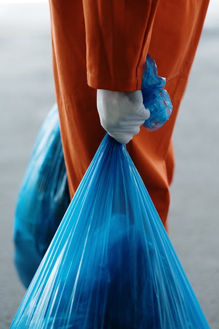 A person wearing a white protective glove is holding open a large white woven polypropylene sack filled with garden waste, including small branches, leaves, and twigs, on an outdoor paved area. The pile of green and brown organic material contains various plant parts with textures ranging from rough and woody to leafy and brittle. A rusty-handled shovel rests against the bag, partially obscured by the waste, indicating active clearing or collection. The surrounding environment appears to be a driveway or garden path, with a person's black footwear visible in the background. The scene suggests an outdoor waste collection or clearance activity, as part of a service such as Rubbish Clearance Highbury involved in disposing of garden or organic waste in accordance with local regulations, offering alternatives to council bulk waste removal.