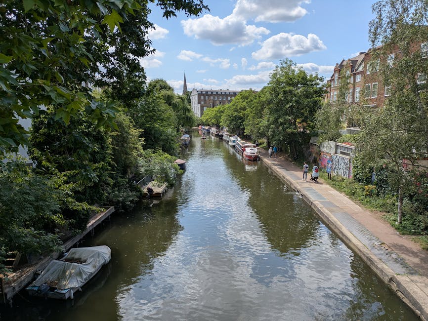 A serene urban canal scene during daytime shows calm, slightly rippled water reflecting the bright blue sky and scattered white clouds. On either side of the canal, there are lush trees with green foliage, some extending over the water's edge, providing natural shade. Along the right side, a narrow paved walkway runs parallel to the water, with a few pedestrians walking and chatting. Several small boats are moored along the banks, some covered with protective tarps. Parked cars are visible on the street behind the trees, and in the background, there are multi-story brick residential buildings with large windows and gabled roofs. A church spire can be seen further in the distance, rising above the tree line. The overall scene depicts a quiet, picturesque area that could benefit from private waste clearance services, like those offered by Rubbish Clearance Highbury, especially for maintaining cleanliness in urban waterways and surrounding environments.