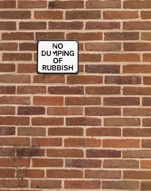 A rectangular metal sign mounted on a red brick wall displaying the message 'NO DUMPING OF RUBBISH.' The sign features a white background with bold black text, with the word 'DUMPING' partially obscured by damage or wear. The bricks in the wall are laid in a traditional running bond pattern, with variations in shades of reddish-brown and some darker bricks interspersed. The mortar between the bricks is light grey. The lighting appears natural, casting a faint shadow of the sign onto the wall. The scene suggests a controlled area for waste disposal, and the sign indicates restrictions on informal rubbish dumping, relevant to services provided by Rubbish Clearance Highbury or similar waste removal companies operating independently of local council rubbish collections.