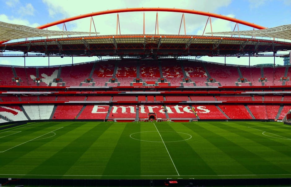 A panoramic view of a football stadium featuring a well-maintained grass pitch with clearly marked white lines and a central circle. The seating area displays rows of red seats with some sections forming patterns or text, with the word 'Emirates' visible prominently in the middle. The stadium has multiple tiers of seating, with some staircases and barriers visible on the sides. The structure is covered by a high, curved roof supported by red framework, extending across the upper tier. The sky above is partly cloudy, and natural daylight illuminates the scene, highlighting the vibrant green of the pitch and the red and white of the seating arrangements, reflecting typical features of a professional football venue, suitable for hosting matches and large-scale sports events.