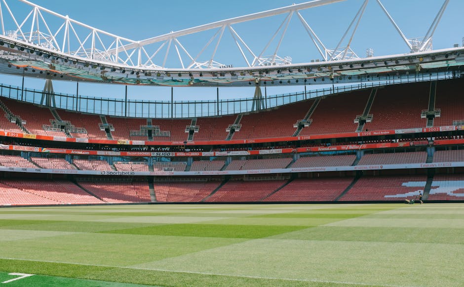 An empty sports stadium with a well-maintained green grass field in the foreground. The seating area features red and pink seats arranged in multiple tiers, with the lower tier partially visible and the upper tier extending around the circumference. A black barrier runs along the edge of the upper seating area, and digital advertising boards are positioned between the tiers displaying various sponsor logos. The stadium's roof structure consists of large white steel trusses forming a geometric pattern above the seating, supporting a transparent or translucent roofing material that allows natural light to filter into the arena. Bright daylight illuminates the scene, highlighting the vibrant colours of the seating and the lush grass, creating a clear and detailed view of the sports venue, which could be associated with private event hosting or alternative recreational use, akin to on-site clearance or maintenance activities typical of rubbish removal or stadium management services. The overall space appears clean, organized, and designed for spectator events, with no visible signs of rubbish or debris within the visible area.