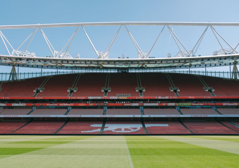 A panoramic view of a football stadium featuring a well-maintained grass pitch with clearly marked white lines and a central circle. The seating area displays rows of red seats with some sections forming patterns or text, with the word 'Emirates' visible prominently in the middle. The stadium has multiple tiers of seating, with some staircases and barriers visible on the sides. The structure is covered by a high, curved roof supported by red framework, extending across the upper tier. The sky above is partly cloudy, and natural daylight illuminates the scene, highlighting the vibrant green of the pitch and the red and white of the seating arrangements, reflecting typical features of a professional football venue, suitable for hosting matches and large-scale sports events.