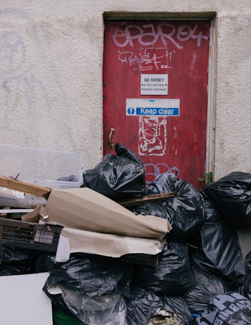 A collection of black rubbish bags piled against a weathered, red metal door embedded in a light-colored, textured wall. The bags are tightly packed and vary in size, with some partially opened revealing contents, resting on a mix of cardboard and discarded packaging materials. The door features multiple graffiti tags in white and light pink spray paint, along with several signs, including a small white sign with black text reading 'NO MONEY,' and a larger blue and white sticker that says 'Keep clear.' The scene appears to be outdoors on a narrow street or alleyway, with the rubbish accumulation indicating a private waste collection or on-site clearance situation handled by Rubbish Clearance Highbury. The overall atmosphere is utilitarian, emphasizing waste disposal in an urban setting, with the visible clutter contrasting against the plain, rough-textured wall and door, supporting the theme of rubbish removal and alternative waste handling services.