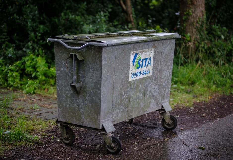 A large, rectangular metal waste bin on four small black caster wheels positioned on an outdoor pavement surface. The bin's surface is weathered with visible dirt, moss, and slight corrosion, giving it a dull, rough grey finish. It features a slightly domed, hinged lid with a handle on the front edge. A faded, partially torn label with the logo and contact number of SITA, a waste management company, is affixed to the side of the bin. The bin is situated at the edge of a paved pathway, with a background of dense green foliage, including trees and bushes, indicating a natural environment or residential area. The lighting is diffuse, suggesting an overcast day, and there are moisture patches on the ground, implying recent rain. This scene exemplifies private or alternative waste handling, typical of rubbish removal services carried out by companies like Rubbish Clearance Highbury, especially in urban or suburban settings where on-site clearance of waste containers is common.