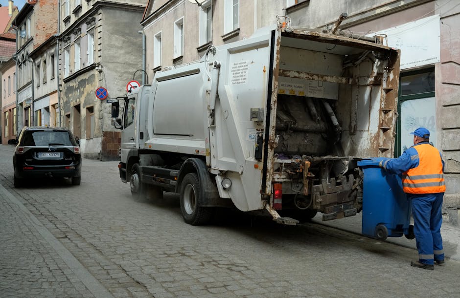 A street scene showing a white industrial rubbish collection truck with an open rear hopper, partially rusted and used for waste disposal, parked on a cobbled road alongside residential buildings with weathered facades. A worker dressed in a blue uniform and high-visibility orange vest is positioned beside the truck, simultaneously holding a blue wheeled bin and preparing to load it into the hopper. The background includes a parked dark grey car and a no-entry traffic sign mounted on a lamppost, with the scene illuminated by natural daylight, highlighting the textures of the street, truck, and clothing. This image exemplifies an independent waste collection activity, aligning with private rubbish removal services offered by companies like Rubbish Clearance Highbury, and showcases on-site rubbish disposal in an urban environment.